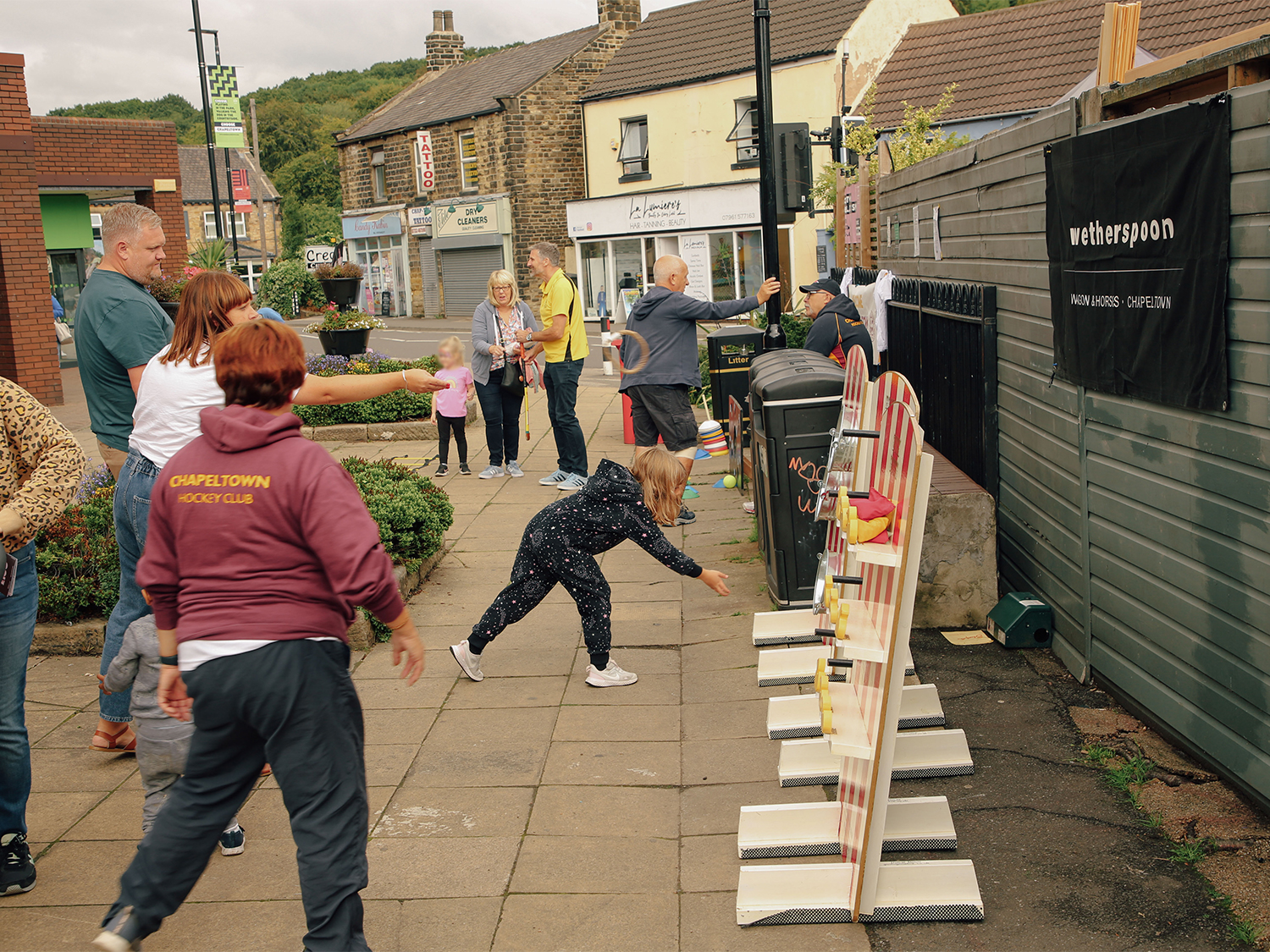 Outdoor community event on a paved area with people playing a game that involves knocking down objects on striped boards. Several participants, including children and adults, are actively engaged, while others watch. Surrounding buildings include stone-fronted shops and a sign for Wetherspoon on a fence. The scene is set in a village or small town with greenery and hills in the background.