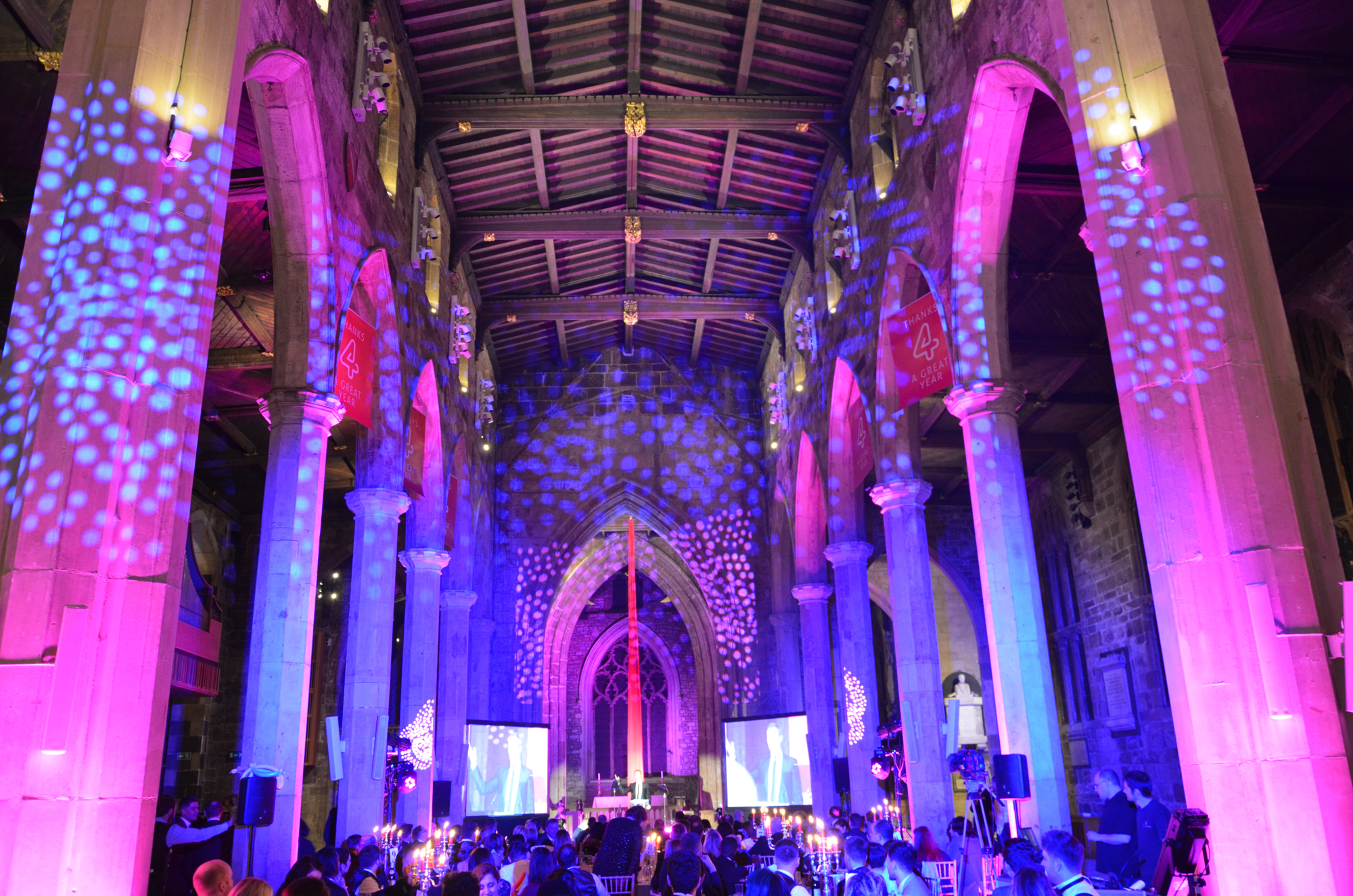 Interior of a large historic cathedral set up for an event, with tall stone columns and high wooden ceiling illuminated by vibrant purple and blue lighting patterns. Two large projection screens flank the central stage area, and guests are seated at tables with glowing decorations.