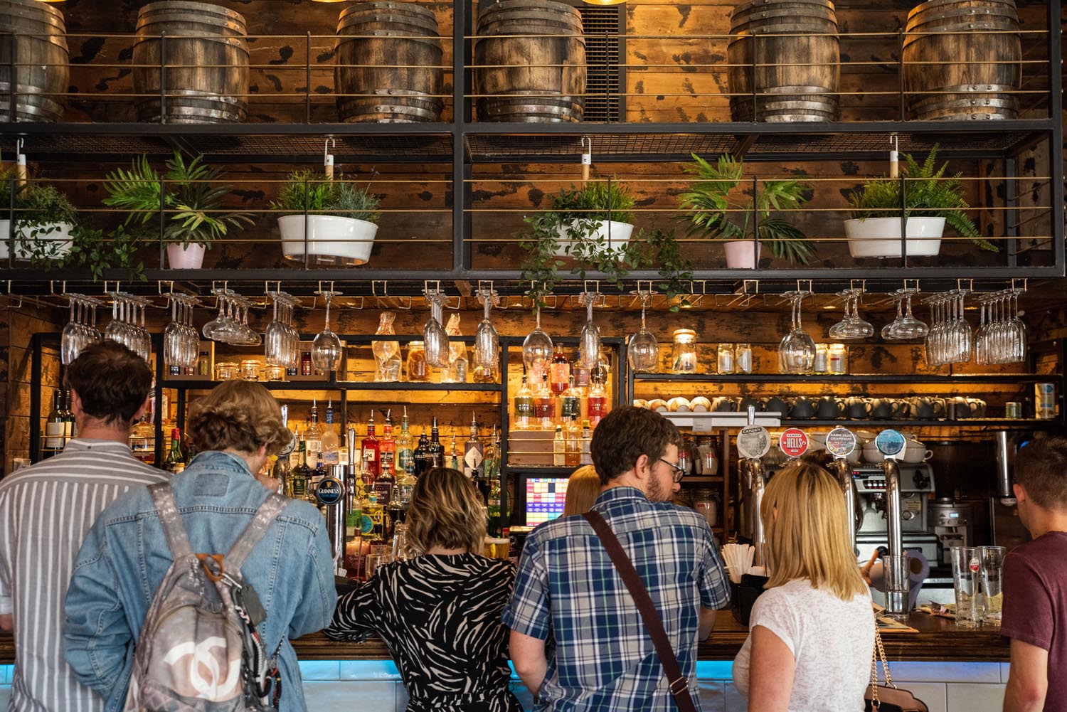 People standing at a bar counter in a rustic-style pub with wooden shelves stocked with bottles and hanging glassware. Above the bar, metal beer kegs and potted plants are displayed on a rack. The warm lighting highlights the wooden textures and industrial decor.