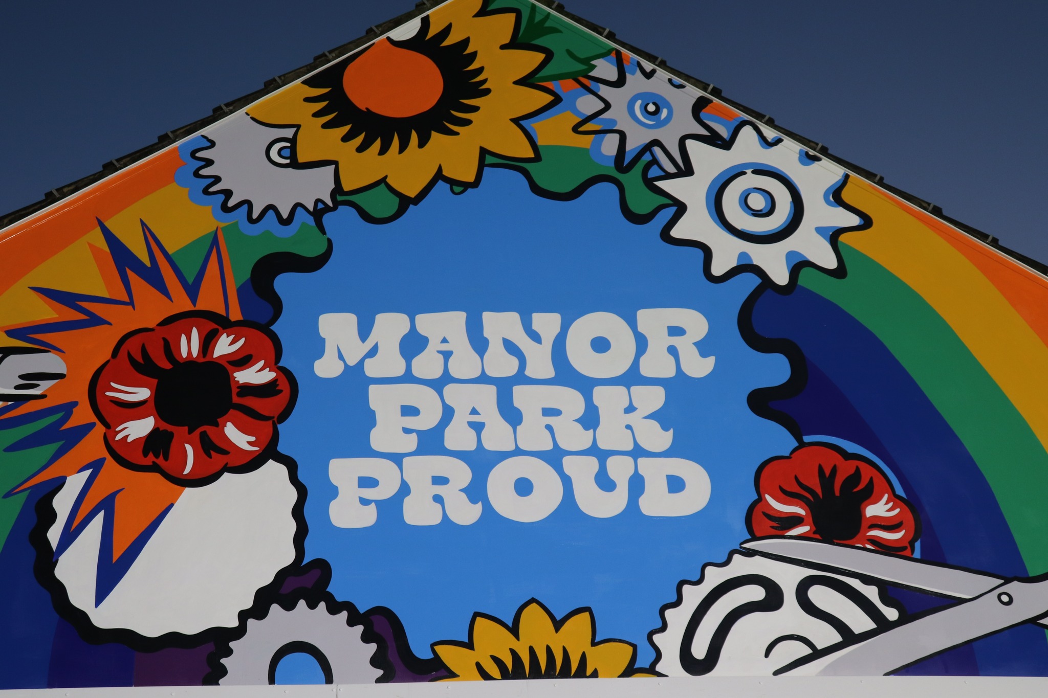 Colorful mural on the side of a building featuring the words ‘Manor Park Proud’ in bold white letters against a blue background. Surrounding the text are vibrant illustrations of sunflowers, red flowers, gears, and a rainbow arching across the top.