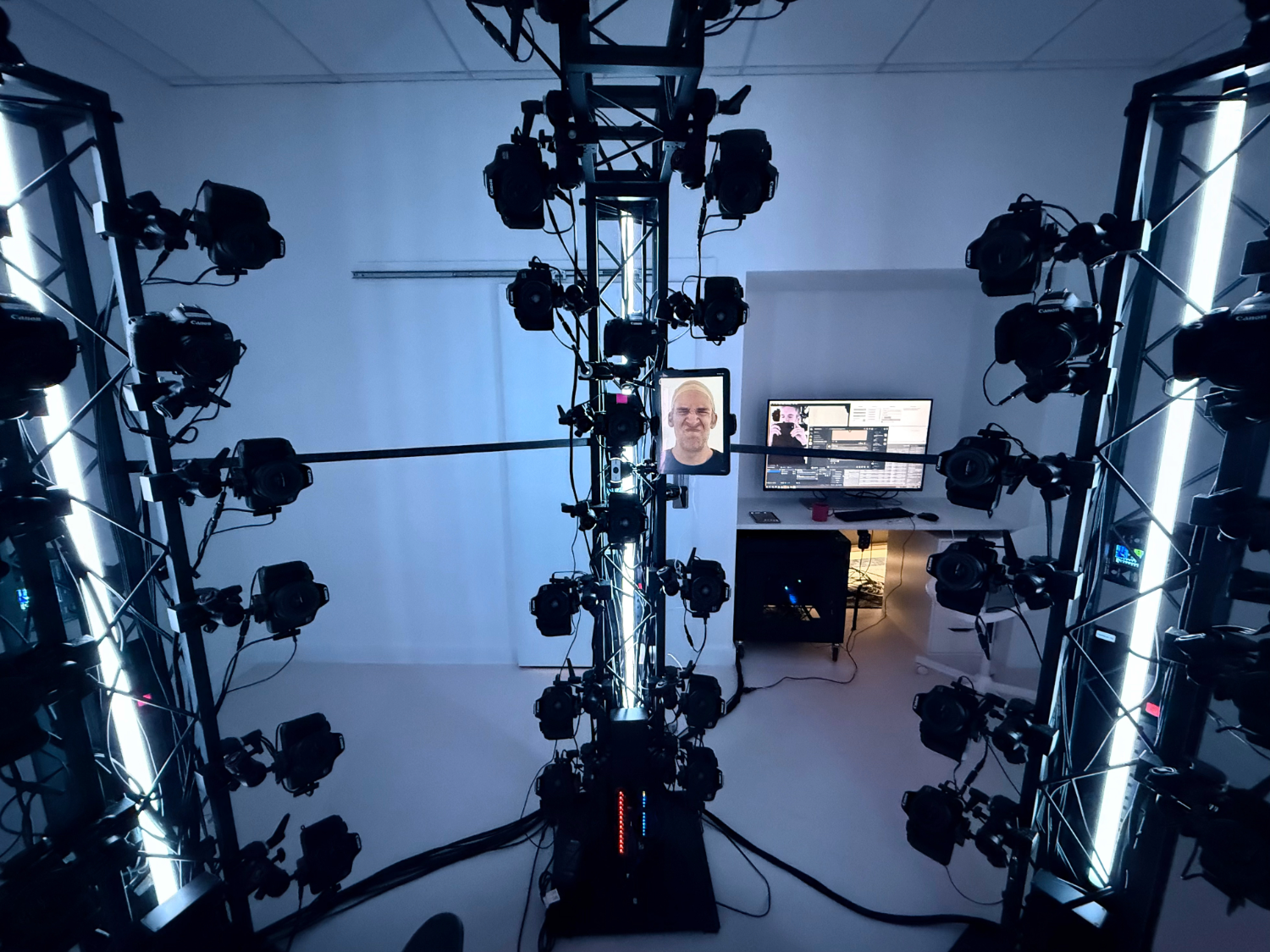 Room with a multi-camera rig arranged on tall metal frames with LED lights, facing a workstation that includes a computer monitor and desk setup.