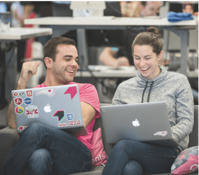 A woman and a man sat next to each other on a sofa. They are working on laptops and laughing.