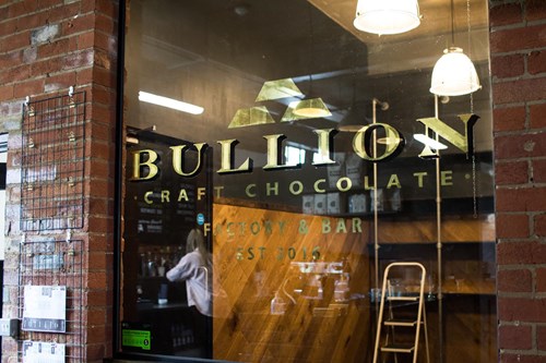 Storefront of Bullion Craft Chocolate, featuring a glass window with the gold Bullion logo and name. Inside, wooden shelves, a ladder, and hanging lights are visible. A person is seen through the window. The building has a red brick exterior.