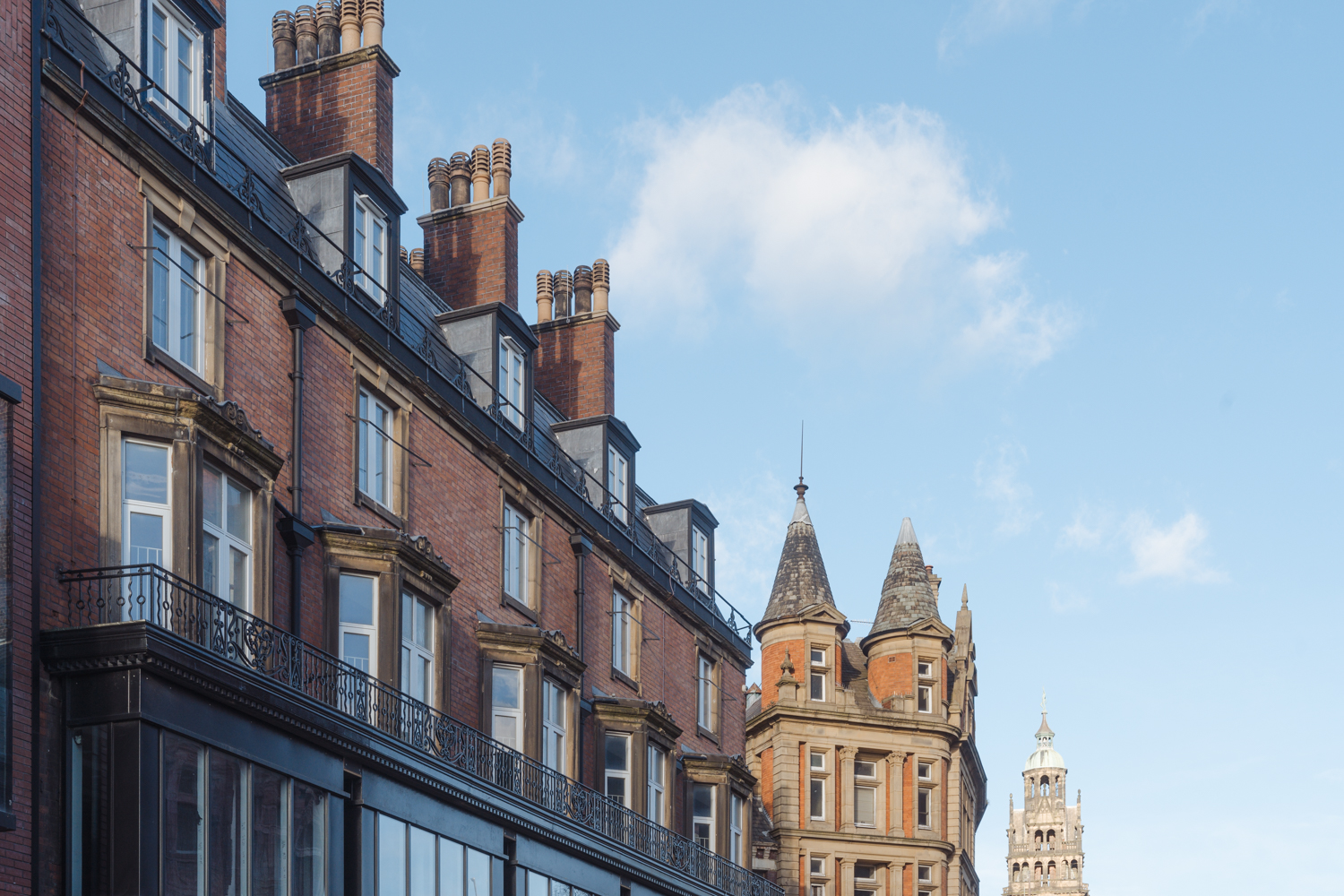 A row of historic red-brick buildings with ornate stone window frames and black wrought-iron balconies, viewed from street level. The architecture features multiple chimneys along the roofline and steeply pitched roofs. In the background, a taller building with decorative spires and a clock tower rises against a clear blue sky with a few scattered clouds.