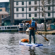 Two people are paddle boarding on the water at Victoria Quays in Sheffield city centre. There are several long boats moored around the edges of the water.