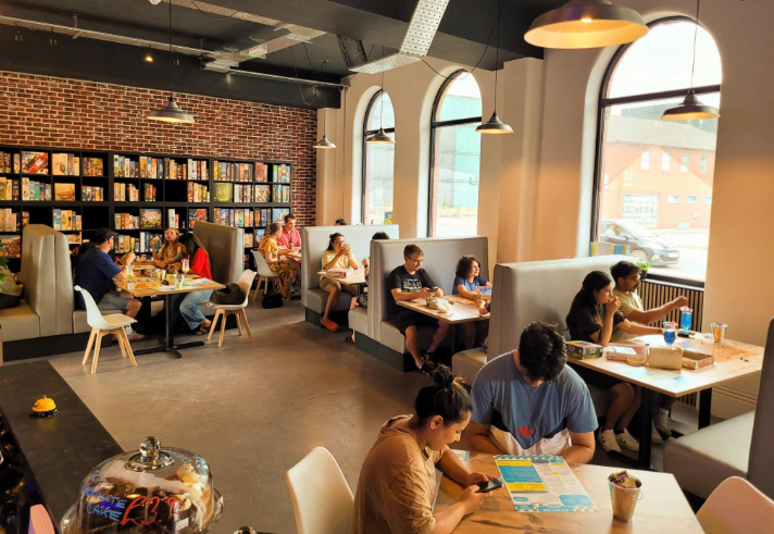 Inside The Dice Box Sheffield, there are tables and booths full of people playing games and drink and eating. The back wall is covered in shelving that is full of boxed games.