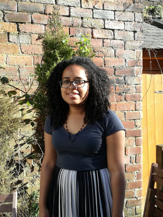 A person standing outdoors in front of a brick wall with some greenery growing against it. The individual is wearing a dark short-sleeve top and a pleated skirt with alternating black, white, and gray panels. There is a wooden chair partially visible in the foreground and a wooden door to the right, suggesting a garden or patio setting.