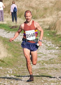 A man fell running, wearing a red vest and blue shorts. There is a race number on his vest.