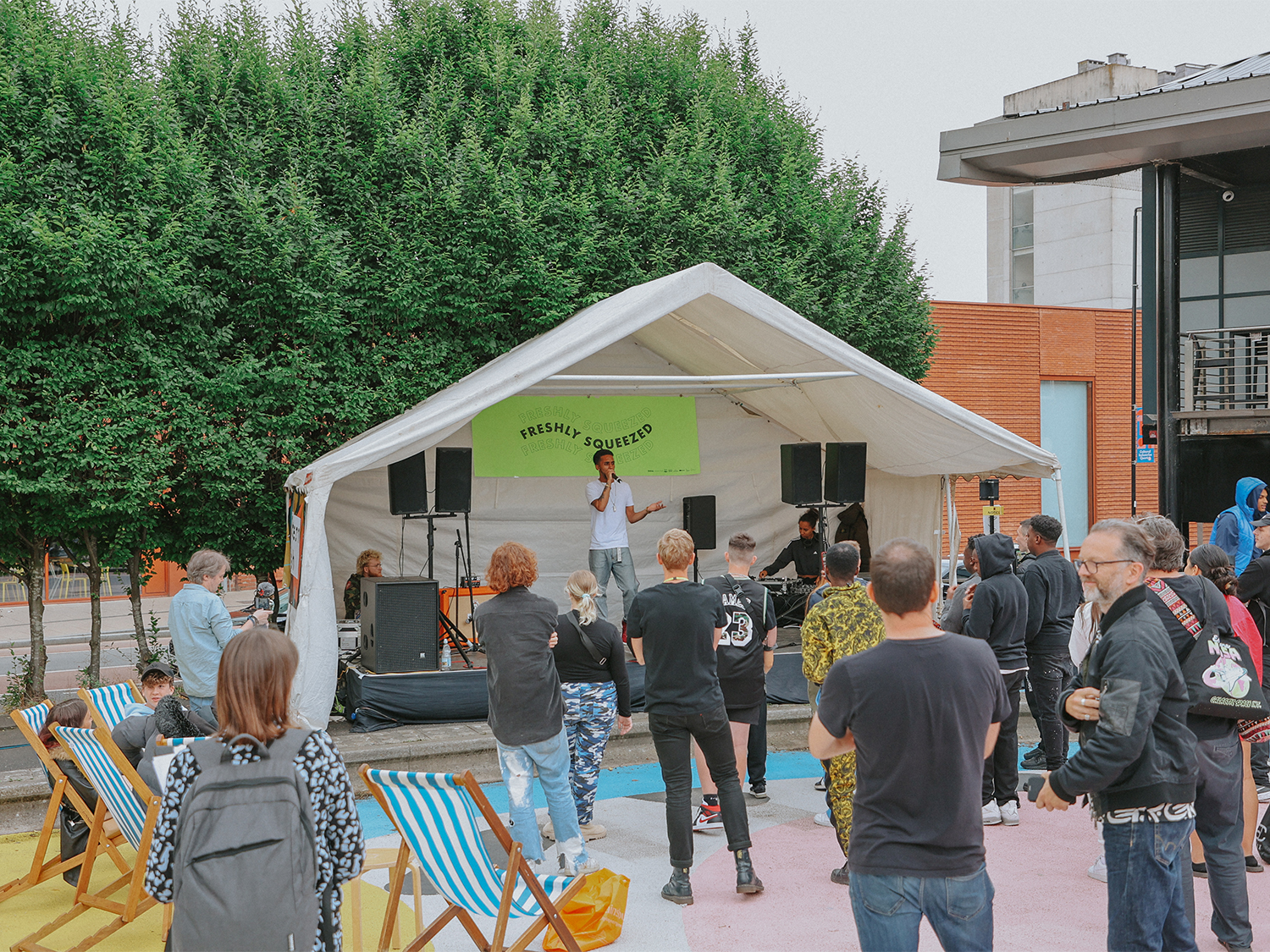Outdoor music performance on a small white tent stage with a green banner reading ‘Freshly Squeezed.’ A performer stands with a microphone while a few musicians sit behind instruments. People are gathered in front of the stage, some standing and others seated on striped deck chairs. The setting includes a large green hedge behind the stage and modern buildings in the background.
