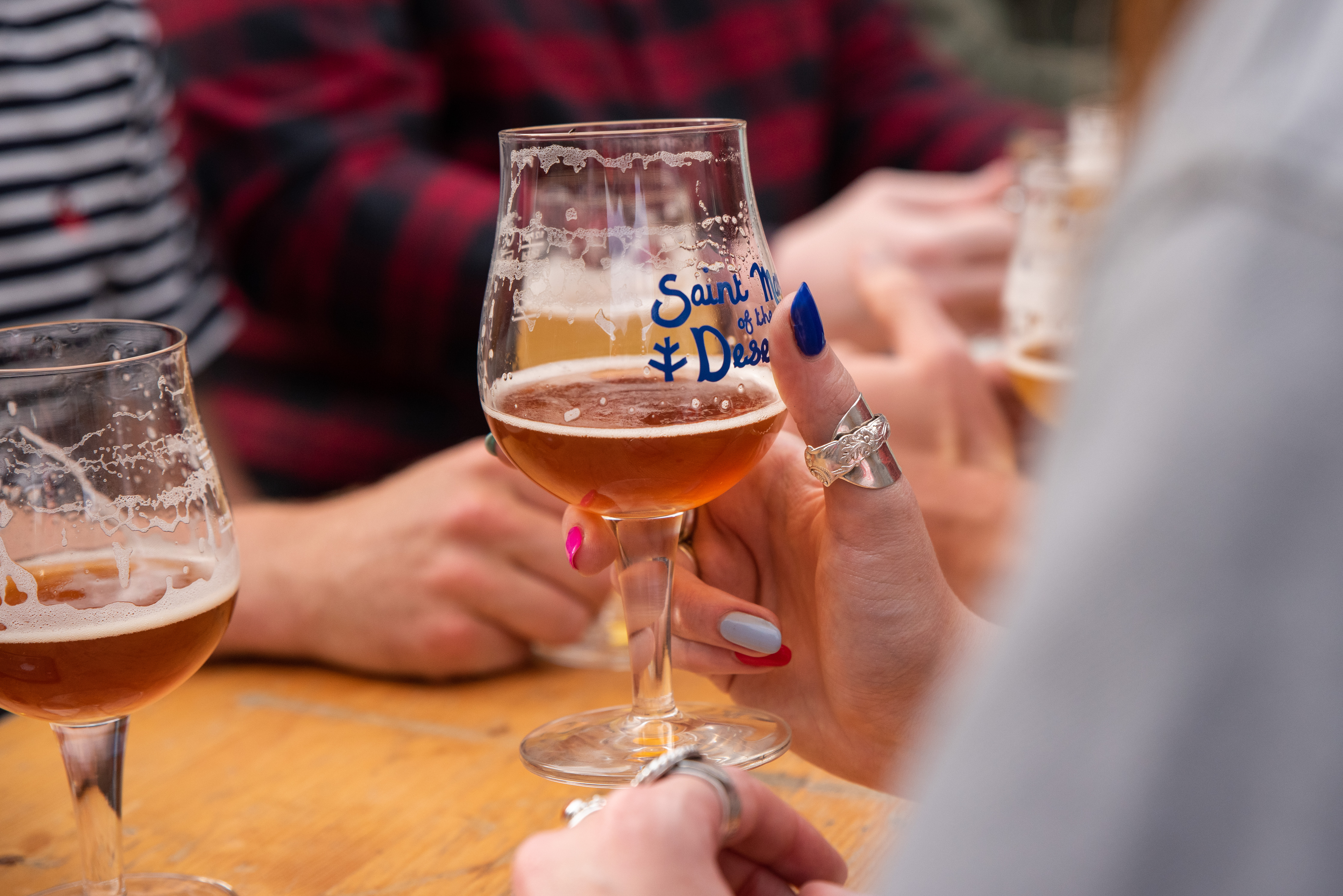 Close-up of several people sitting around a wooden table with multiple glasses of craft beer. One hand in the foreground holds a glass labeled ‘Saint Mars of the Desert.’ Other glasses with similar labels and varying shades of beer are visible, along with a notepad and pen on the table.