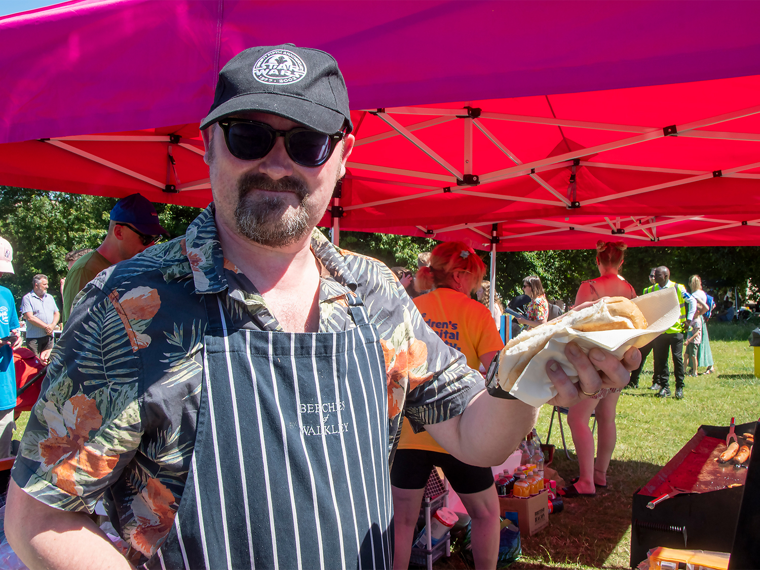 Person wearing a floral shirt and striped apron stands under a bright pink canopy at an outdoor event, holding a wrapped food item. Behind them, other people are gathered near tables and stalls, with snacks and drinks visible. The scene is set on grass with trees in the background, suggesting a summer fair or community festival.