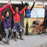 People cheering with their arms in the air, outside a building. There are several mountain bike lent up against the building.