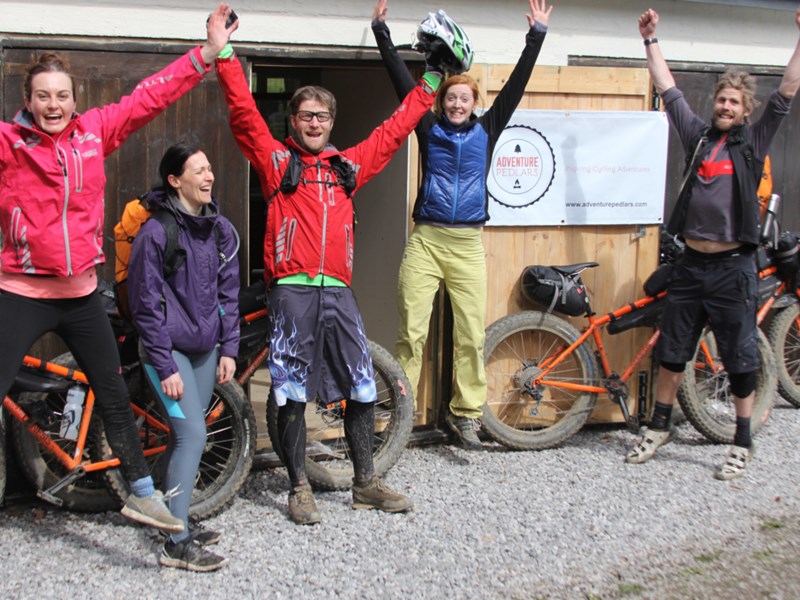 People cheering with their arms in the air, outside a building. There are several mountain bike lent up against the building.