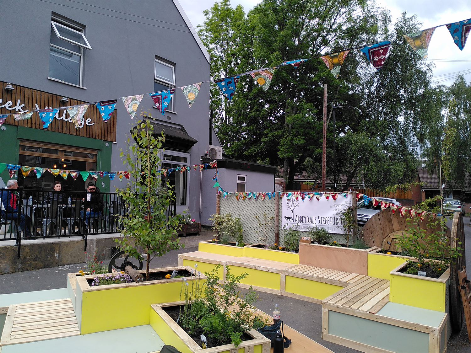 Outdoor seating area in front of a café decorated with colorful triangular bunting. The space features bright yellow and light blue wooden planters with small trees and plants, integrated benches, and a banner reading “Abbeydale Street” in the background. A few people are seated at tables near the café entrance, and large green trees are visible behind the building.
