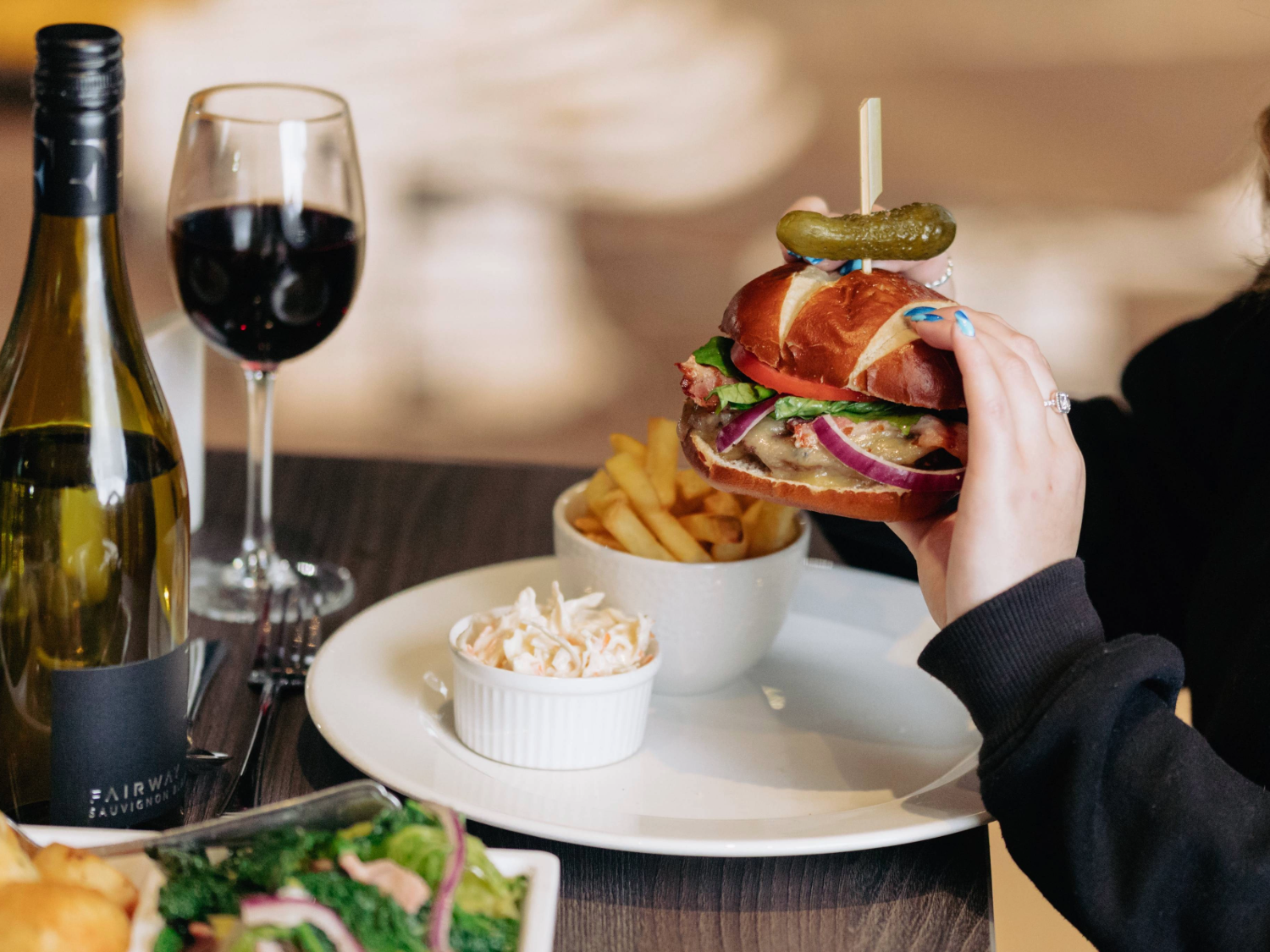 A fancy burger plus chips and a side salad and a bottle of wine laid out on a table. A woman is picking up the burger.