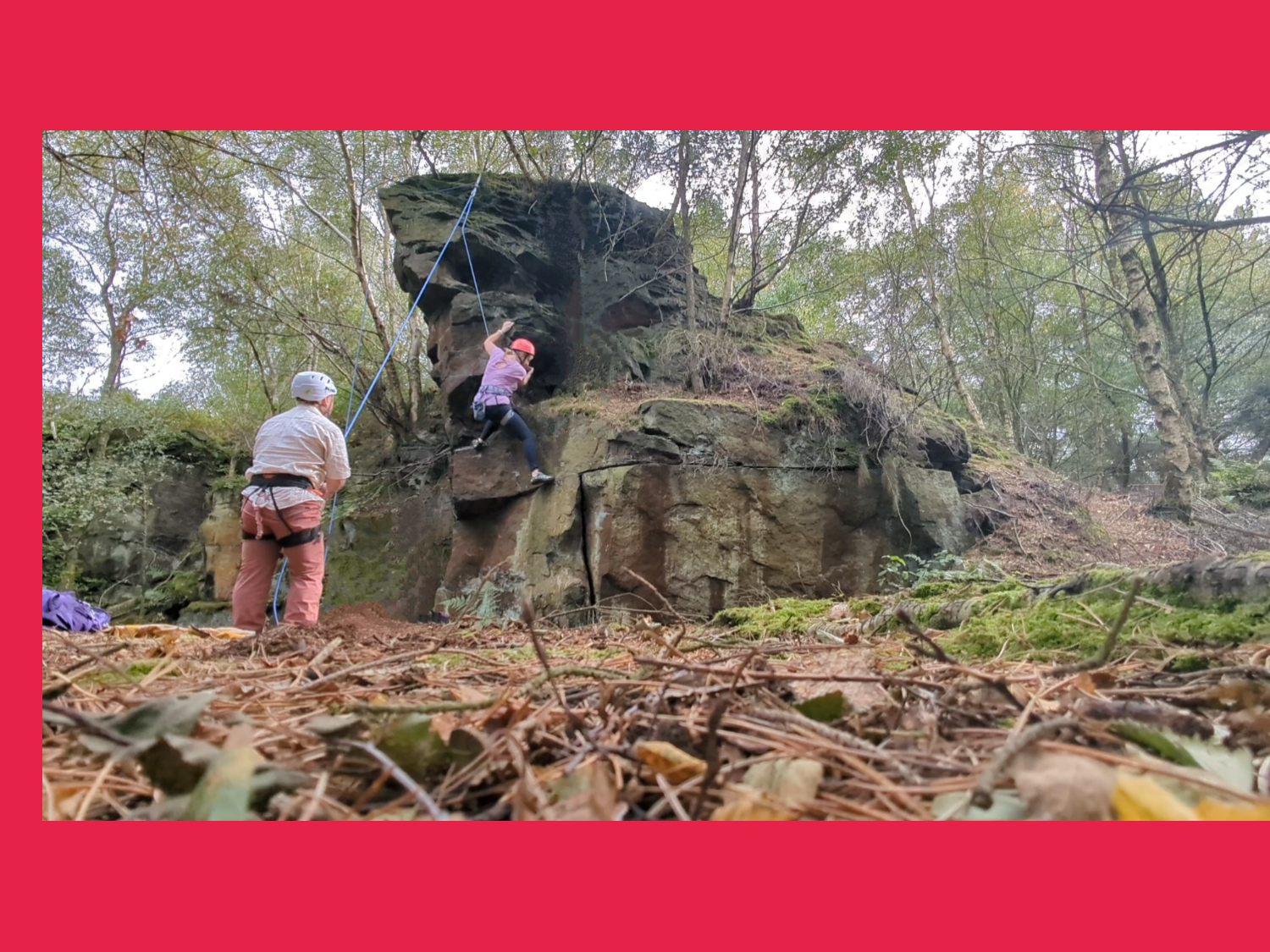 Two people bouldering at a rocky outcrop in the woods.