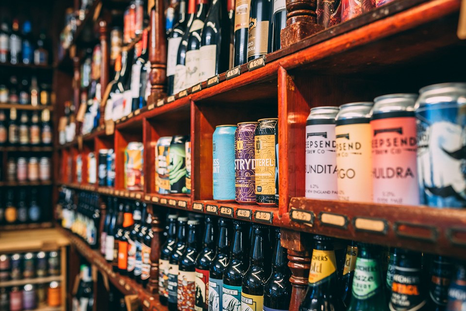 A store shelf filled with a wide variety of beers, including colorful cans and neatly arranged bottles. Additional shelves in the background display more options, suggesting a large selection.