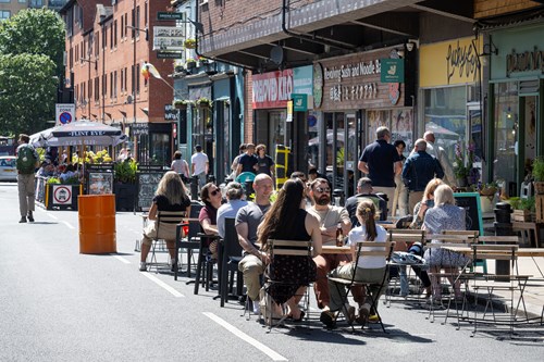 People are seated at outdoor tables on a closed-off city street, enjoying food and drinks in sunny weather. The street is lined with shops and restaurants, including signs for a noodle bar and a café. A bright orange barrel and potted plants decorate the area, while pedestrians walk along the street in the background. The scene conveys a lively, social atmosphere in an urban setting.