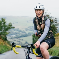 Female cyclist laughing as she takes a break, countryside can be seen in the background.