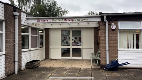 Entrance to a single-story building with a sign above the double doors that reads “Welcome to Sheffield Mencap & Gateway.” The exterior features a mix of white and brown brick walls, large windows, and a paved area in front. A wooden bench and a blue wheelbarrow are positioned near the doorway, and trees are visible in the background under an overcast sky.