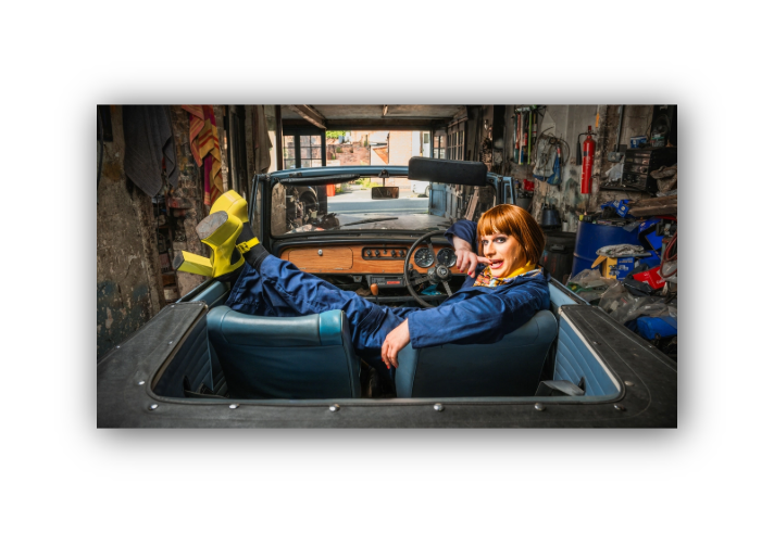 A man, in make-up and a wig and wearing blue overalls and bright yellow platform shoes, lounges in an open top-car in a small garage that is full of all sorts of tools, spare-parts and cloths and rags.