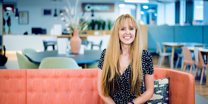 A woman, sat on a sofa, looking straight at the camera and smiling. Behind her is an open-plan office in soft focus.