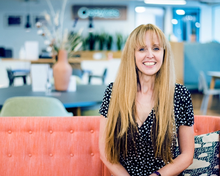 A woman, sat on a sofa, looking straight at the camera and smiling. Behind her is an open-plan office in soft focus.