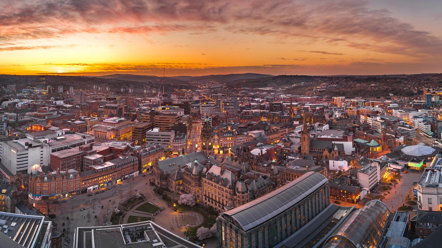 Panoramic view of Sheffield city at sunset, with warm orange and pink hues in the sky. The foreground features historic buildings, including the ornate Sheffield Town Hall, surrounded by modern structures and glass-roofed buildings. Streets and squares are visible below, with city lights beginning to glow. Rolling hills form the backdrop on the horizon.