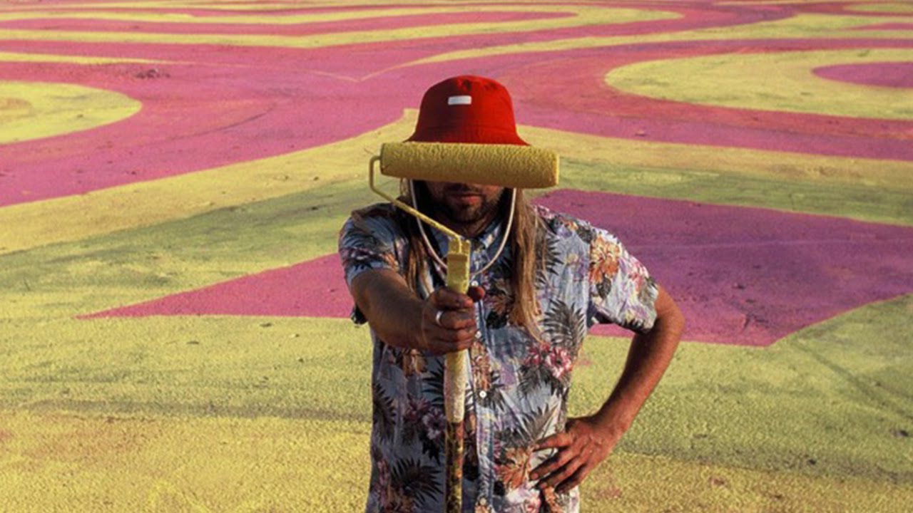 Street artist INSA holds a paint roller in front of his eyes to hide his identity with one of his artworks of swirling yellow and pink shapes in the background 