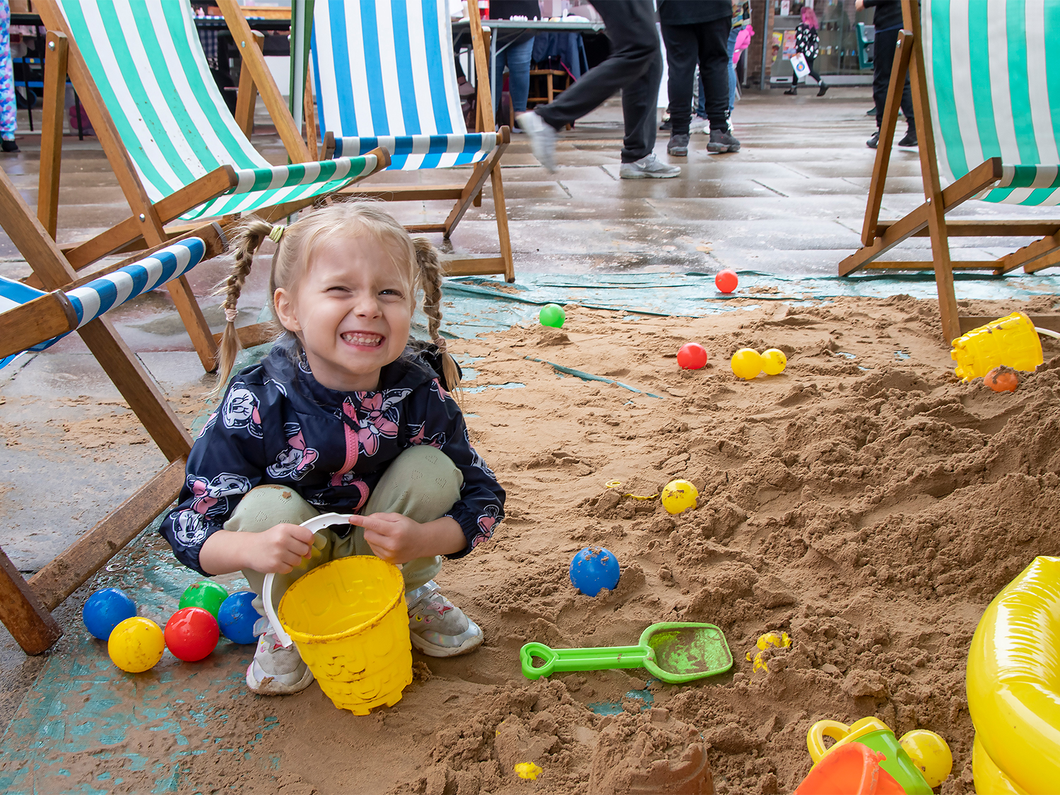 Child playing in an indoor sandpit surrounded by colorful plastic toys, including a yellow bucket, green spade, and small balls. Several striped deck chairs are positioned around the sand area, and people can be seen walking in the background.