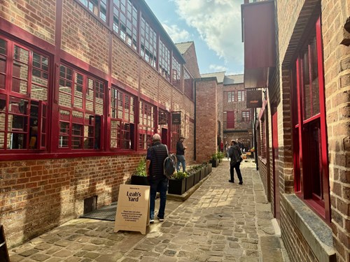Cobblestone alleyway between historic red-brick buildings with large red-framed windows. Several people are walking along the passage, which is lined with rectangular planters containing greenery. A wooden sign on the left reads ‘Leah’s Yard.’ The scene is lit by natural daylight under a partly cloudy sky.