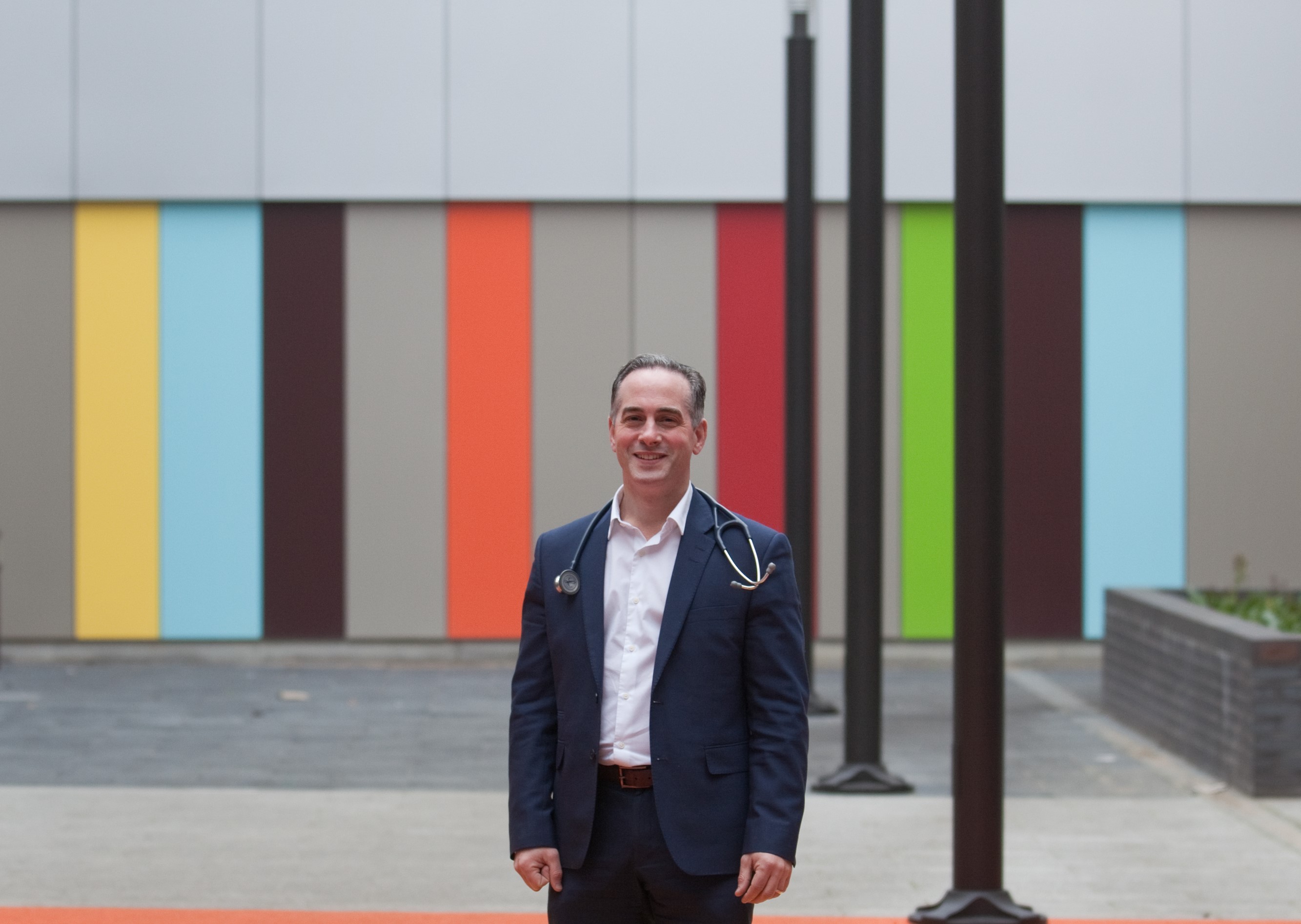 Person wearing a dark blue suit and white shirt with a stethoscope draped around the neck, standing outdoors in front of a modern building facade featuring vertical panels in bright colours including orange, yellow, green, blue, and brown. The area has a paved surface and tall black poles.