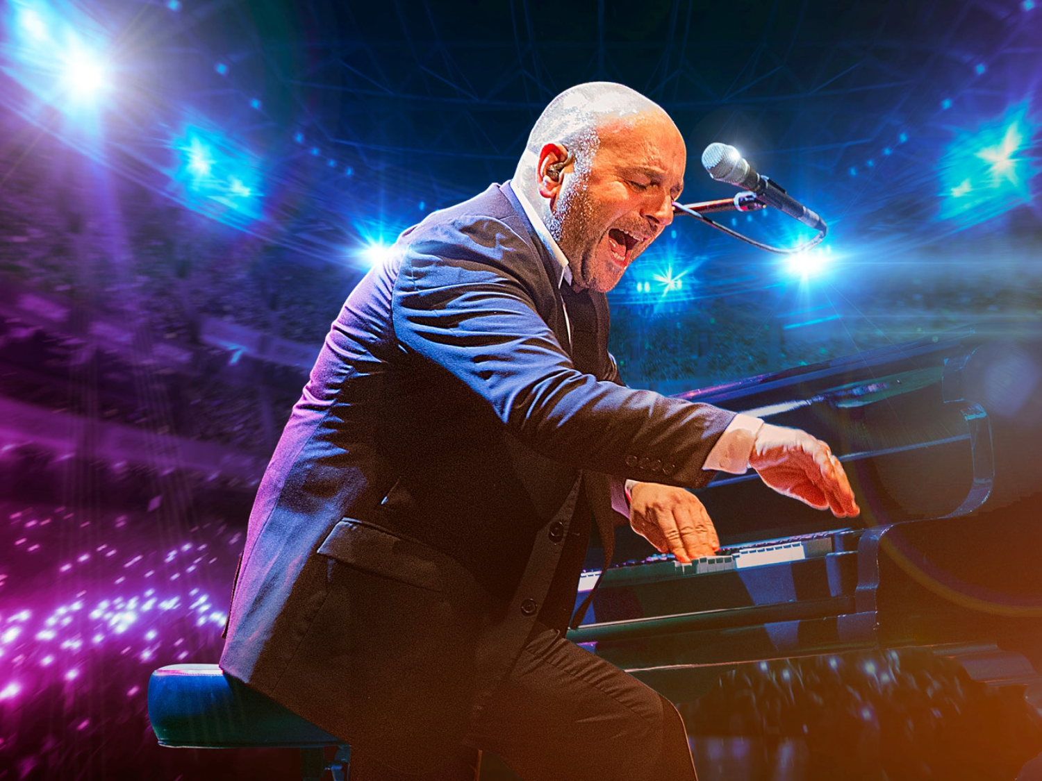 A performer in a dark suit is seated at a grand piano on a brightly lit stage. The musician is leaning forward with one arm extended toward the keys, while a microphone is positioned above the piano. The background shows vivid blue and purple stage lights and a large audience area illuminated with glowing lights.