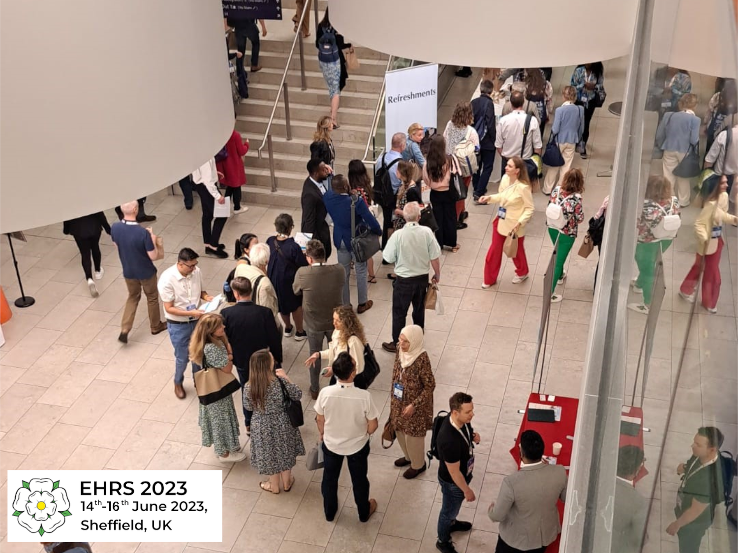 Busy conference venue with attendees networking and walking around a spacious atrium. People are gathered near a staircase and refreshment area, with tables set up for registration or materials. A sign reads “Refreshments,” and a logo in the bottom corner shows “EHRS 2023, 14th–16th June 2023, Sheffield, UK” with a white rose emblem.