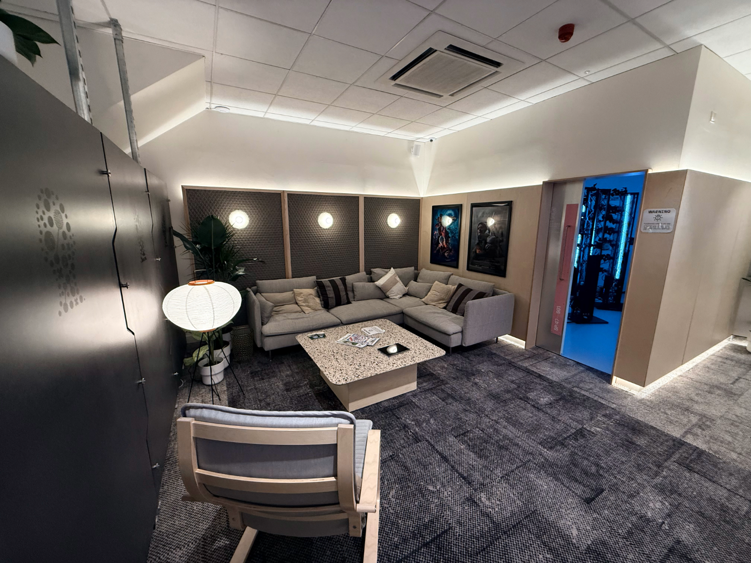 Modern lounge area with a large L-shaped sofa, cushioned seating, and a low coffee table in the center. The room features soft wall lighting, framed artwork, a decorative floor lamp, and a doorway leading to a blue-lit adjacent space.