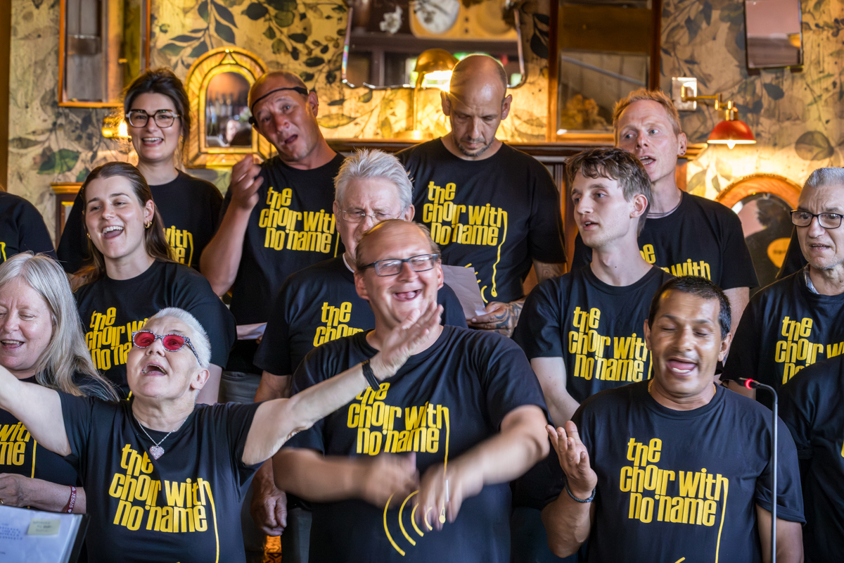 Members of The Choir With No Name singing together in black t-shirts with yellow text, standing in front of a decorative wall with mirrors and lights.