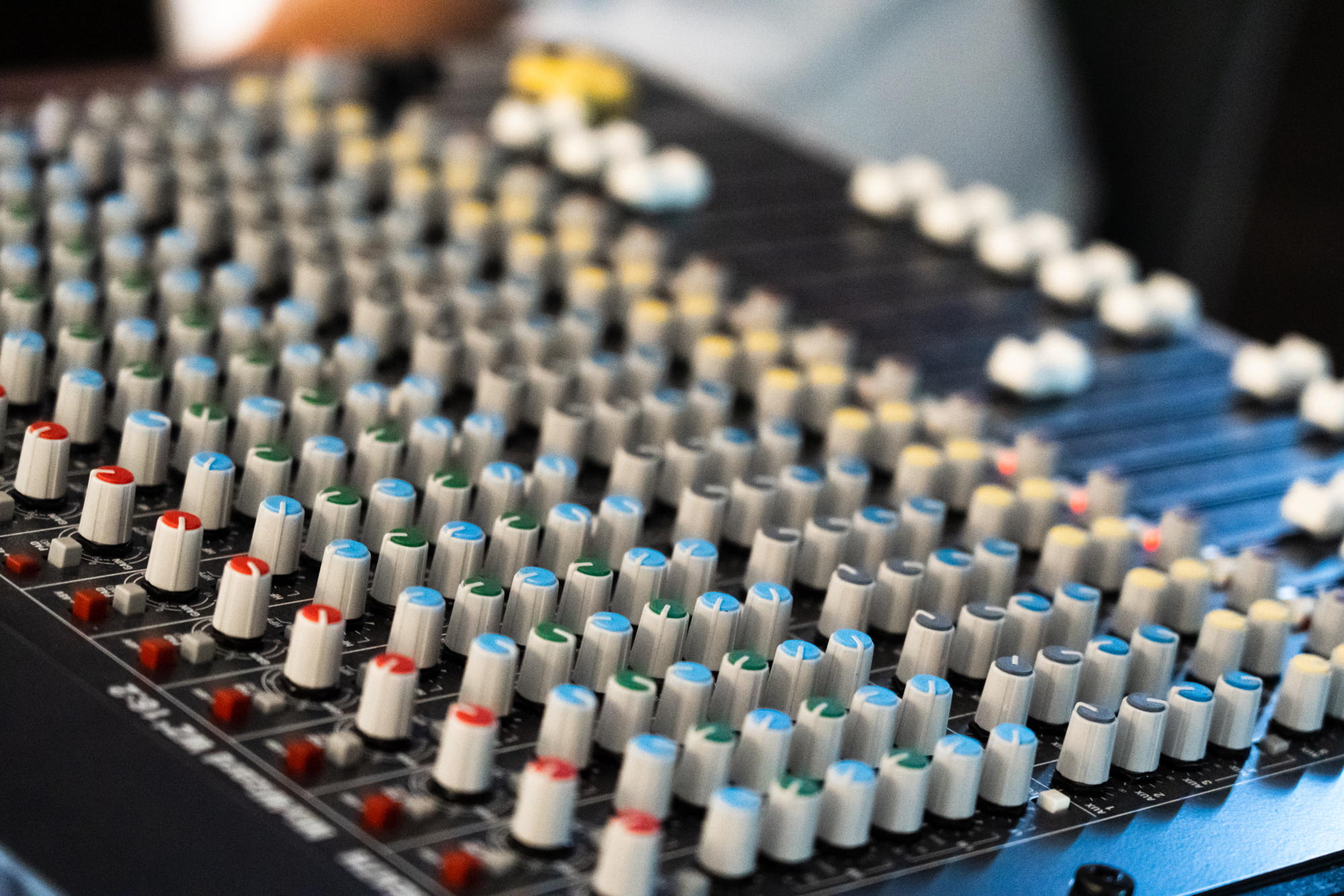 Close-up view of an audio mixing console with numerous knobs and sliders arranged in rows, featuring different coloured caps for channel control.