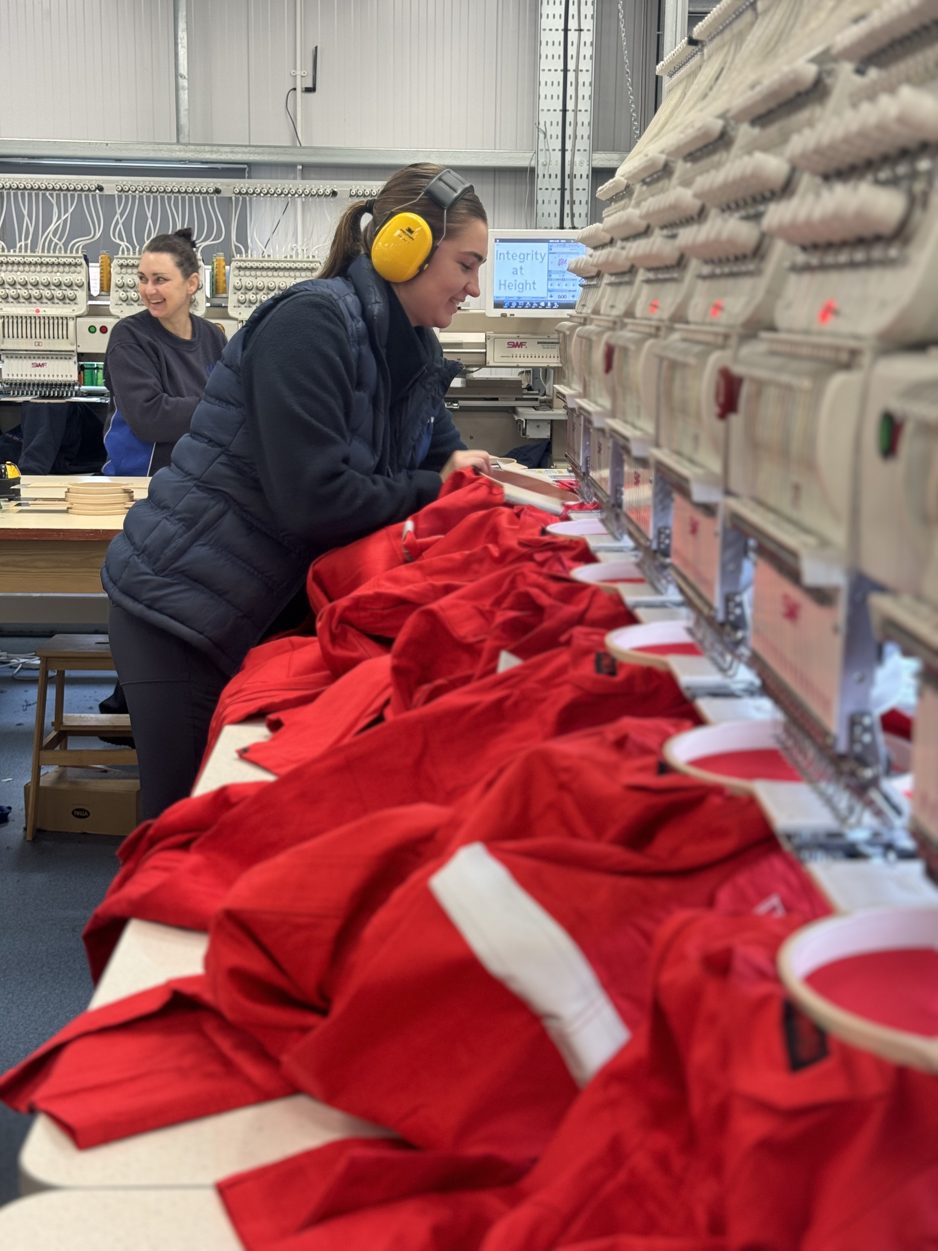 Person wearing ear protection and a padded jacket operating a row of embroidery machines in a workshop. Bright red garments are laid out on the machines for stitching. Another person is visible in the background, and the workspace includes tables and equipment under bright indoor lighting.