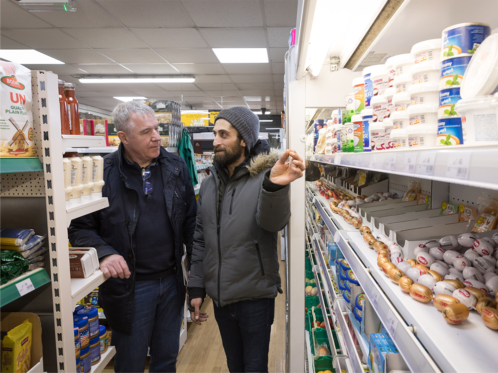 The inside of a food shop, with the owner and a customer standing between rows of shelves. 