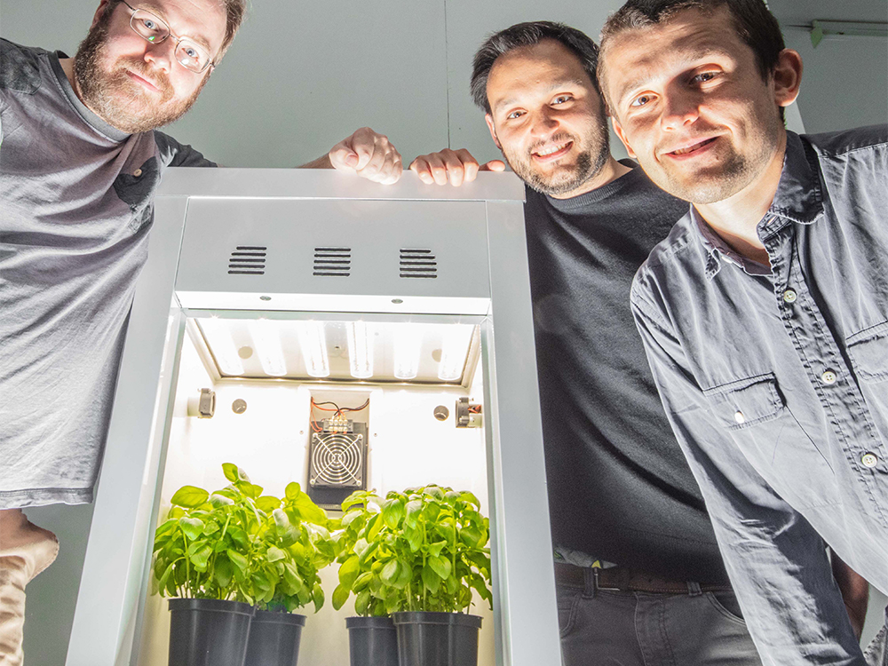 Three people stand around a white, illuminated growth chamber containing two potted plants with green leaves. The chamber features a fan and lighting system at the top. All three individuals are smiling and looking at the camera