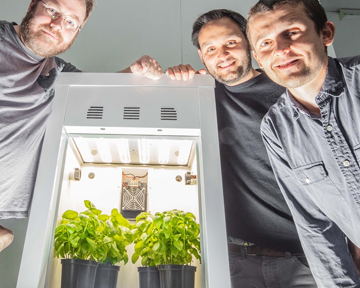 Three people stand around a white, illuminated growth chamber containing two potted plants with green leaves. The chamber features a fan and lighting system at the top. All three individuals are smiling and looking at the camera