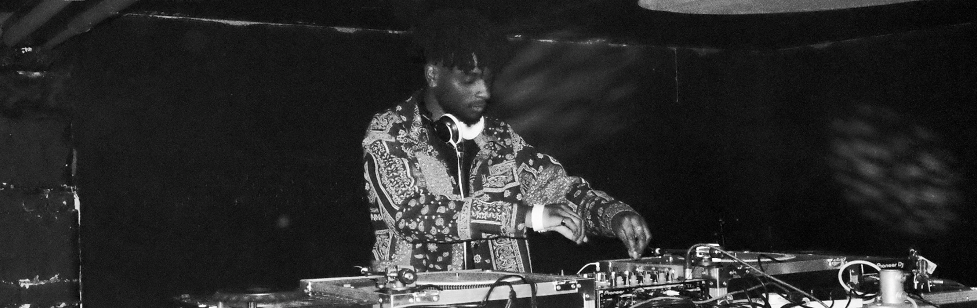 A black and white image of a DJ standing behind two record decks and a mixer, playing music.