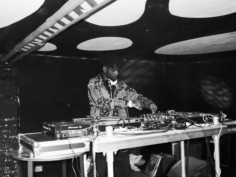 A black and white image of a DJ standing behind two record decks and a mixer, playing music.