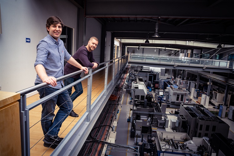 Two people standing on an upper-level walkway inside a large industrial facility, leaning on a metal railing and overlooking rows of advanced manufacturing machines and equipment below. The space features high ceilings, exposed beams, and bright overhead lighting, creating a modern factory environment.