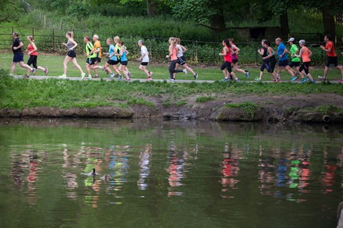 A group of people on a park run, running along a path that runs beside a pond.