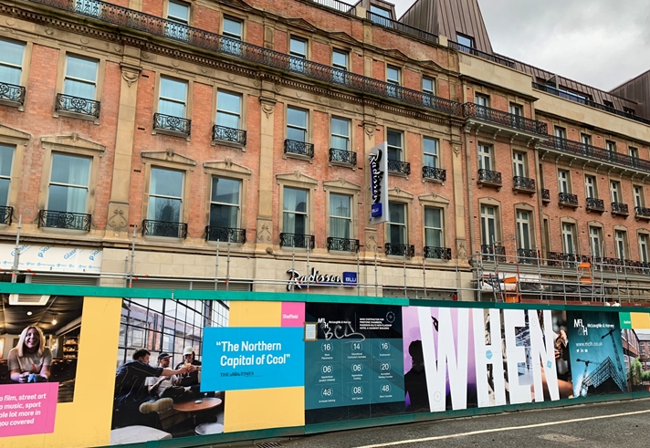 Street view of a historic building with a Radisson Blu hotel sign, featuring ornate windows and black wrought-iron balconies. In front of the building is a colorful construction hoarding with promotional graphics and text, including phrases like “The Northern Capital of Cool” and “WHEN.” The hoarding displays images of people indoors, event details, and branding elements. Scaffolding is visible along the lower part of the building, and the sky is overcast.