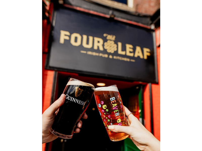 A pint of beer and a pint of Guinness being clinked together under The Four Leaf Irish Bar & Kitchen's sign.