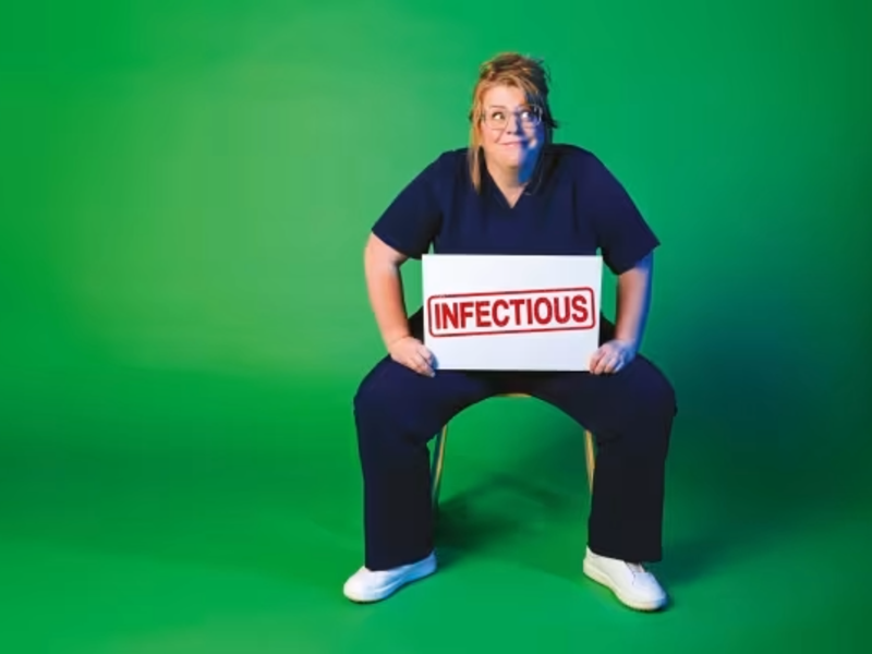 Nurse Georgie Carroll sits on a chair against a green background. She is wearing scrubs and holds a large sign that say 'Infectious'.