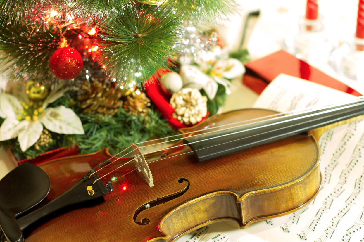 A violin on a table covered in sheet music and a Christmas ornament off to one side.