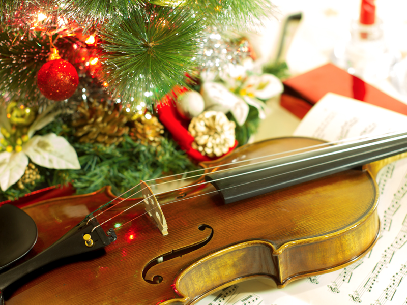 A violin on a table covered in sheet music and a Christmas ornament off to one side.